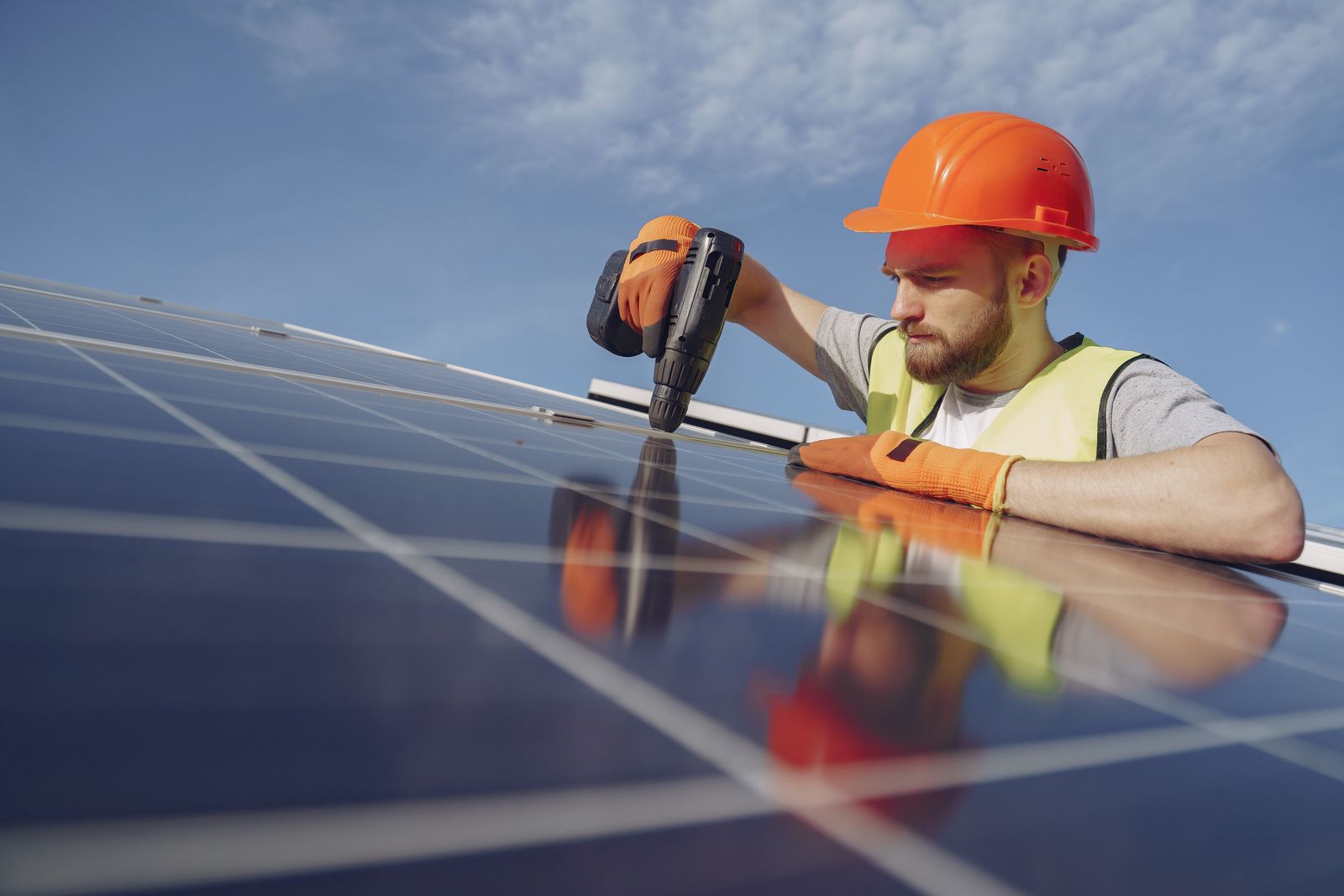 Elektryk w pomarańczowym kasku i kamizelce odblaskowej wkręca wkręt w ramę panelu fotowoltaicznego na dachu