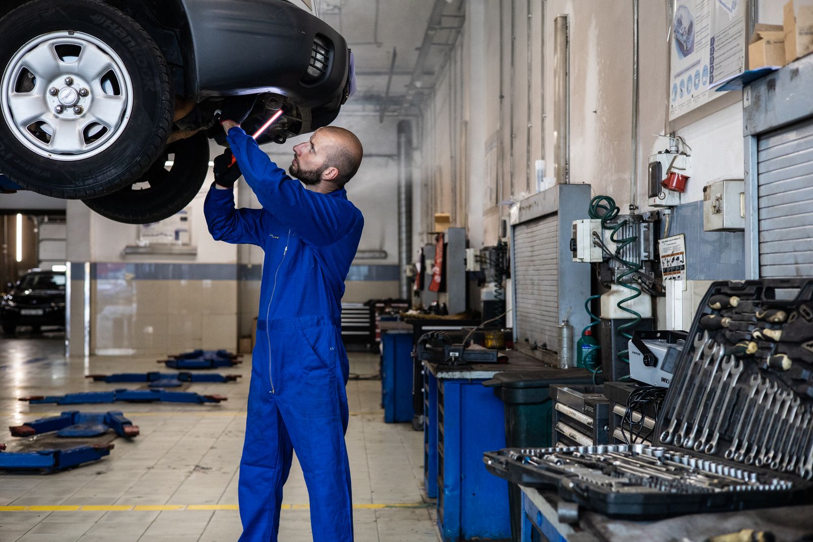 Mechanik w granatowym kombinezonie przy aucie podniesionym na podnośniku, z wiertarko-wkrętarką w dłoni i wózkiem z narzędziami obok