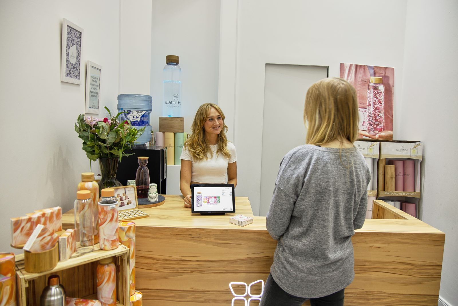 Jasny blat z drewna przy recepcji salonu, donica i półki z produktami; za lady stoi recepcjonistka rozmawiająca z klientką.