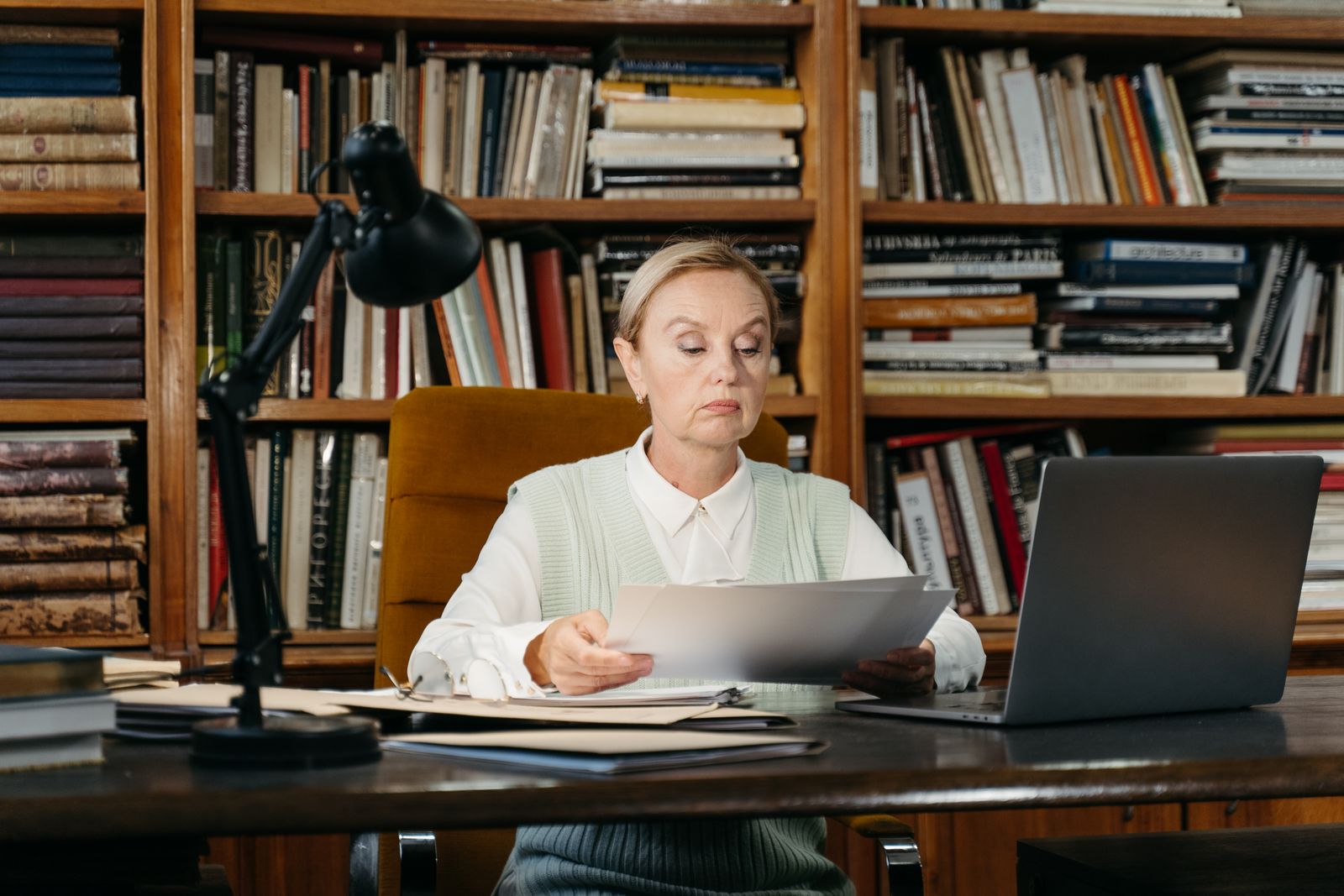 Sekretarz stowarzyszenia czyta dokument przy biurku w niewielkim gabinecie otoczonym regałami z książkami; obok stoi laptop i lampa biurkowa